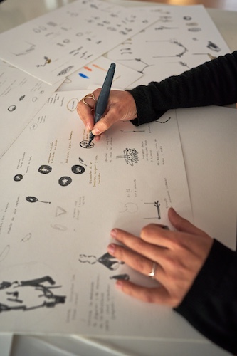High angle of crop anonymous talented female designer drawing sketch of different accessories on paper in jewelry workshop